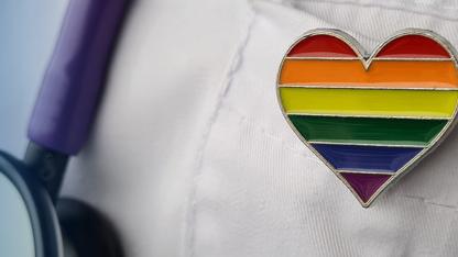 Anstecker in Form eines Herzens in Regenbogenfarben steckt an einem Arztkittel, daneben ein Stetoskop.